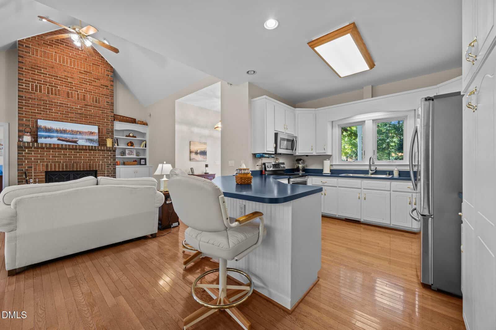 Spacious kitchen with white cabinets, blue countertops, and stainless steel appliances open to a living room with a brick fireplace, vaulted ceiling, and hardwood floors.