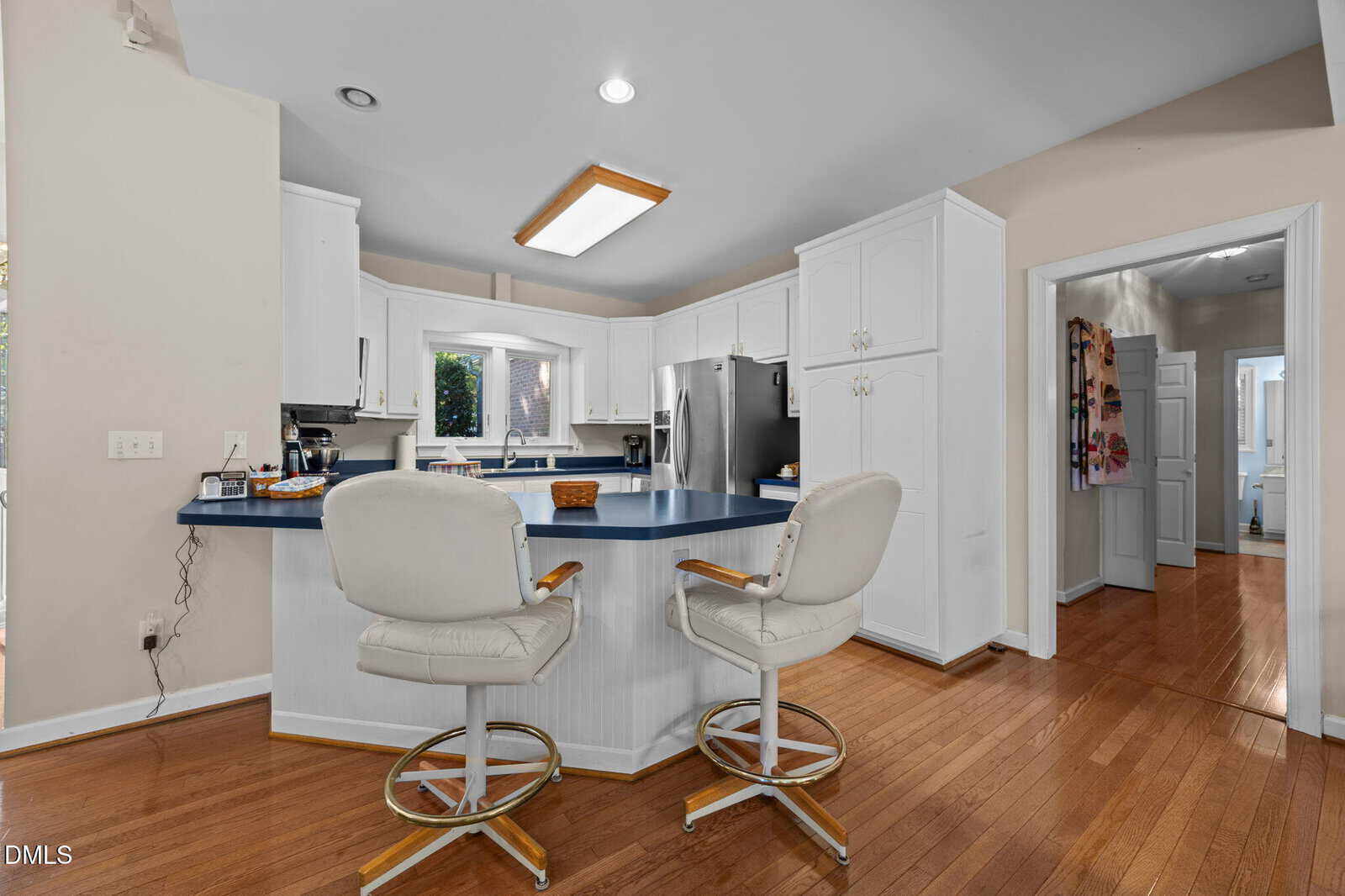 Bright kitchen with white cabinets, blue countertops, and two white swivel barstools on a hardwood floor. Stainless steel appliances and a hallway are visible in the background.