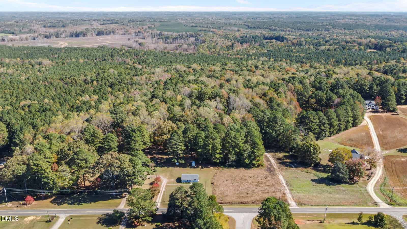 Aerial view of a rural landscape with a single house centered on a large plot, surrounded by dense forest, open fields, a few dirt roads, and a paved road in the foreground.