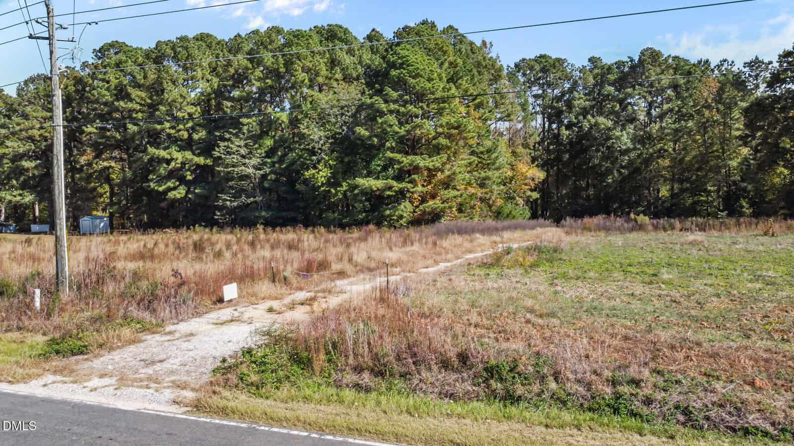 A gravel driveway runs from a paved road through an overgrown field with dry grass and weeds. Power lines cross overhead, and dense trees line the background under a blue sky.