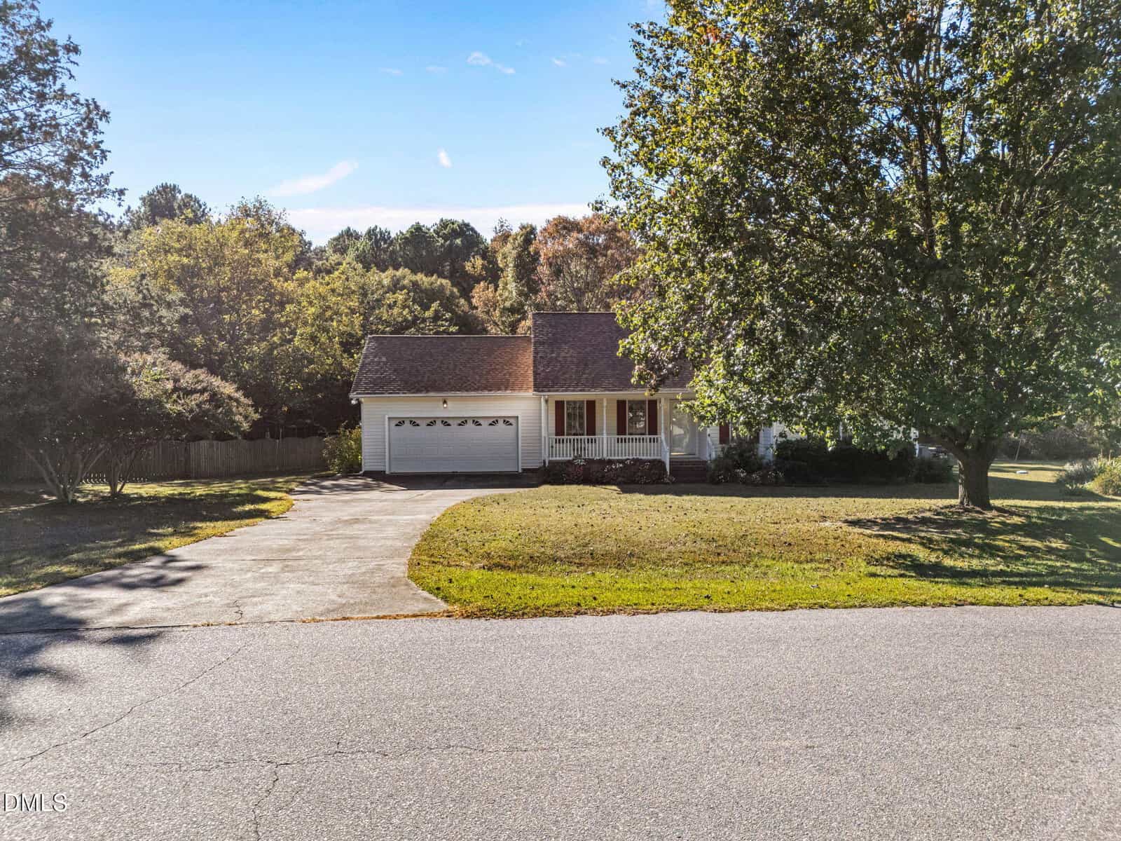 Single-story house with a front porch, attached two-car garage, and driveway. Large front lawn with mature trees, bordered by a wooden fence and surrounded by greenery under a clear blue sky.