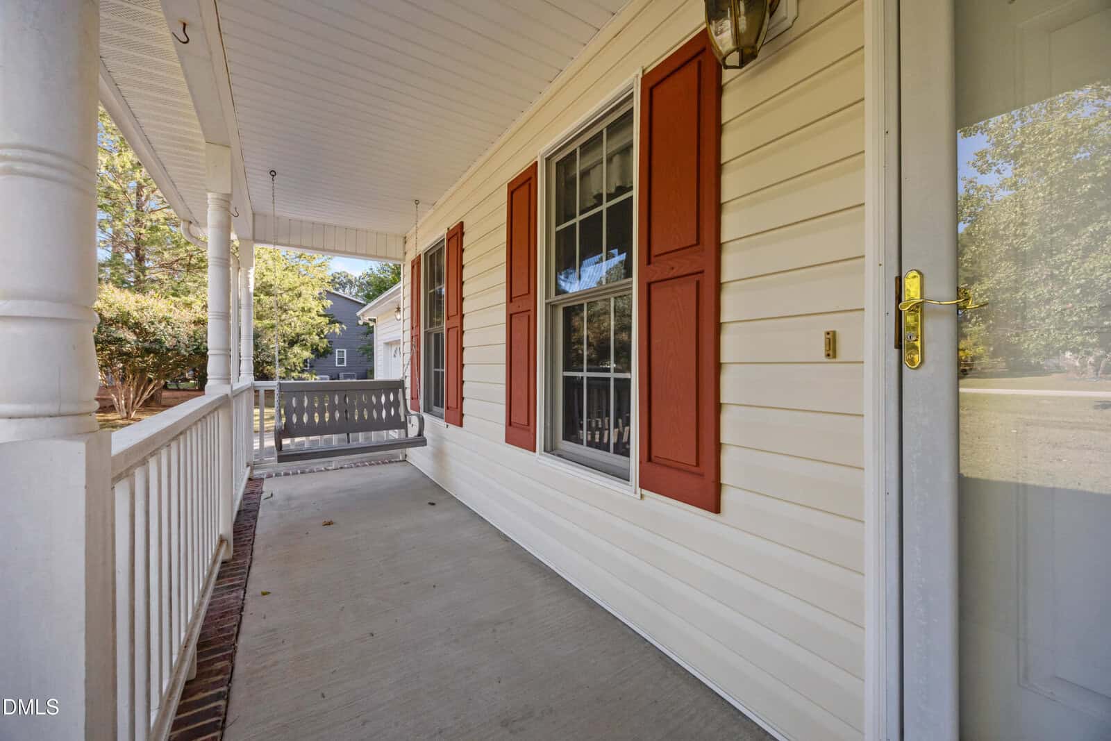 A covered front porch with a white railing, beige siding, two windows with red shutters, a glass storm door, a porch light, and a gray porch swing. Trees and a neighboring house are visible in the background.
