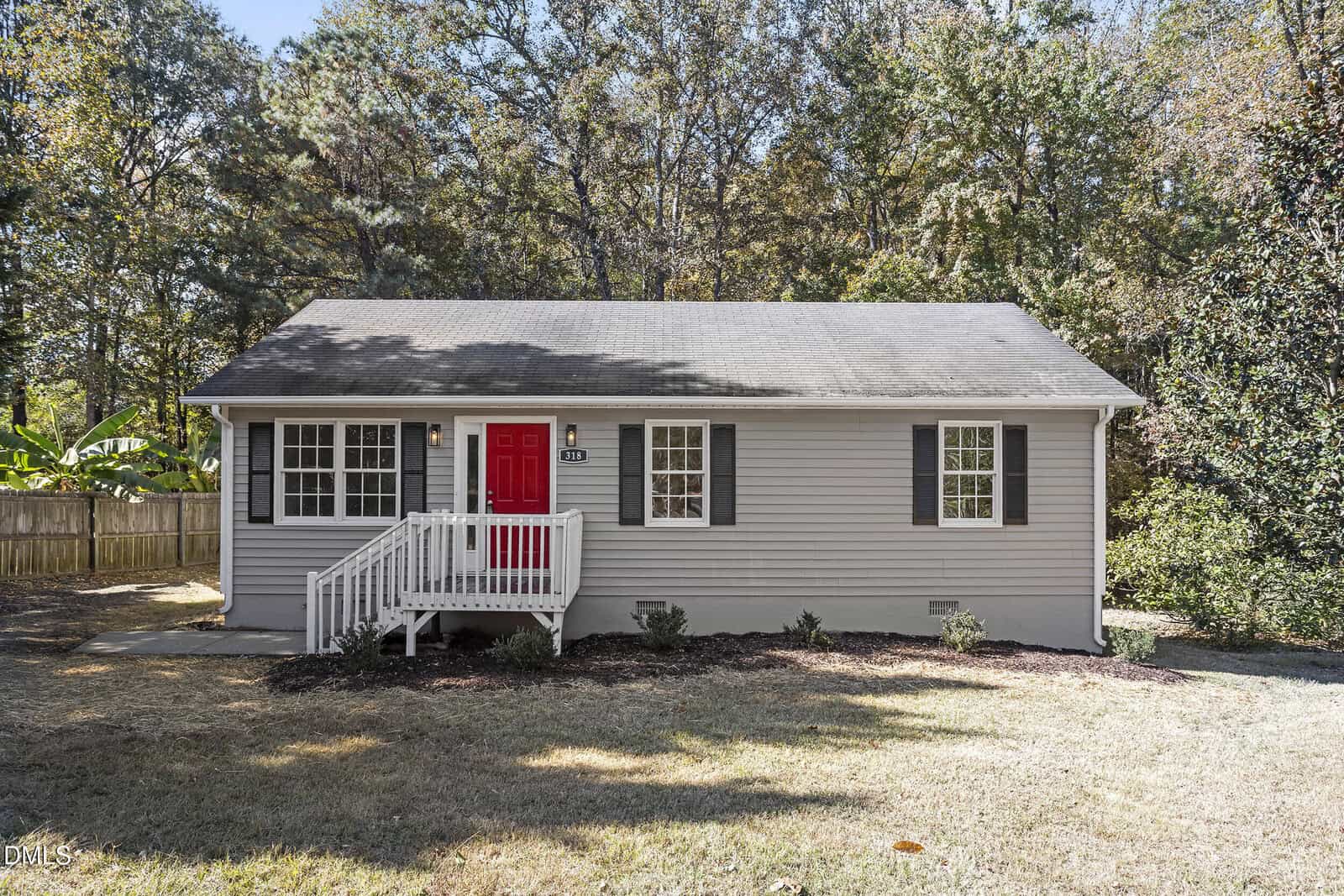 Nestled at 318 Apple Street in Creedmoor, this small, single-story house features light gray siding, black shutters, and a charming red front door. A white porch railing and a few small shrubs complete the inviting look. Trees and a wooden fence sit behind.