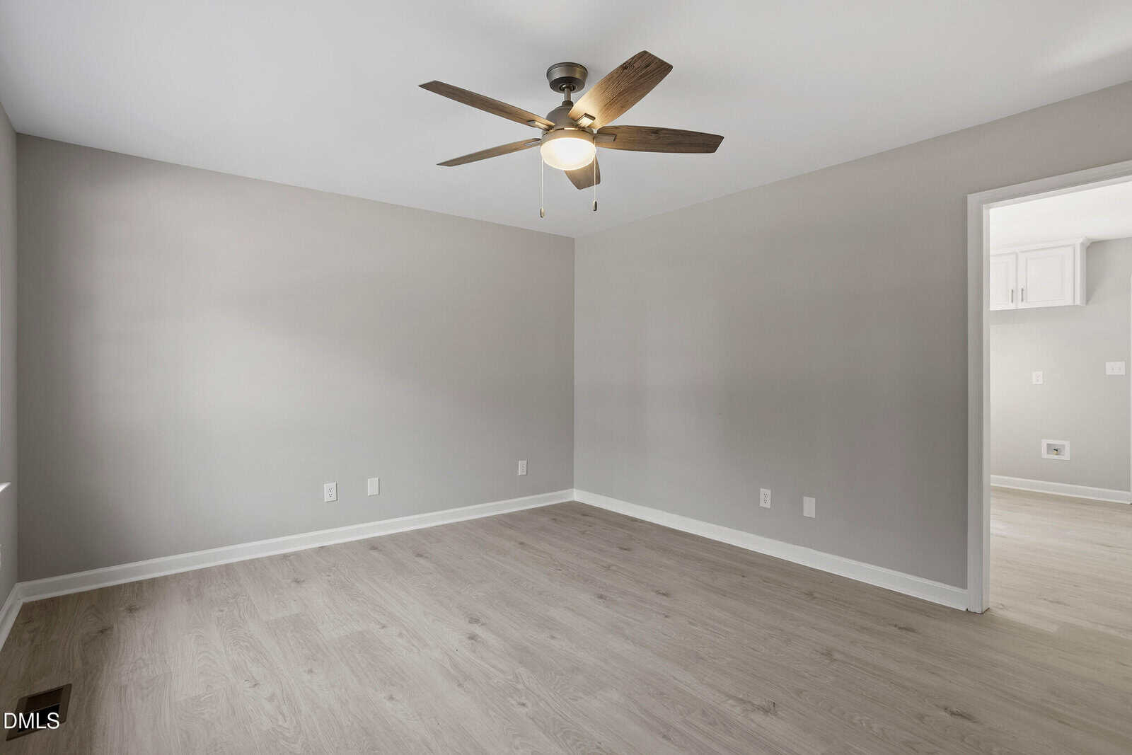 Empty room with light wood flooring, light gray walls, a ceiling fan with wooden blades and a light, and an open doorway leading to another room with similar flooring and partial view of white cabinets.
