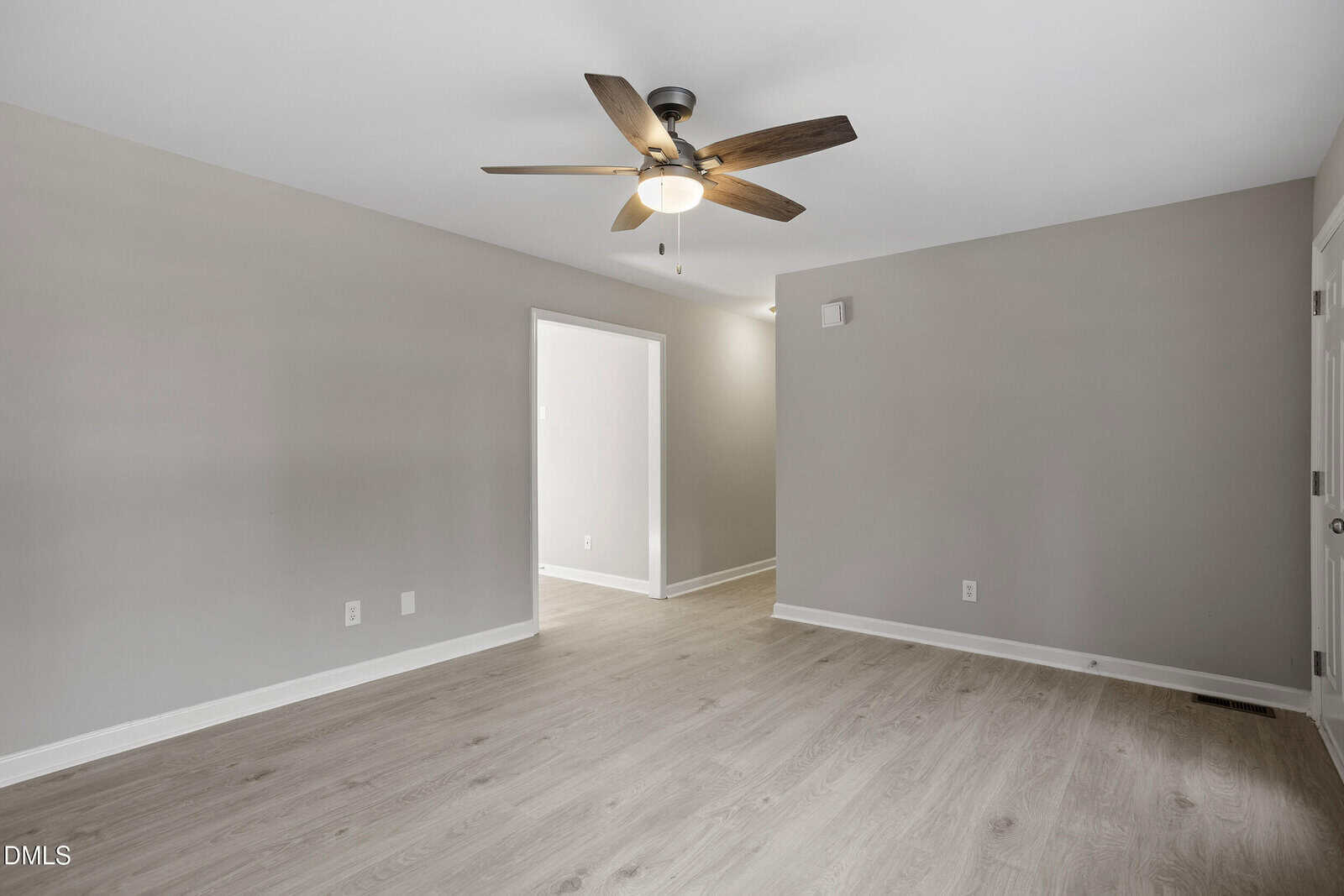 A clean, empty room with light wood flooring, beige walls, a ceiling fan with lights, and an open doorway leading to another area. There are multiple electrical outlets on the walls.