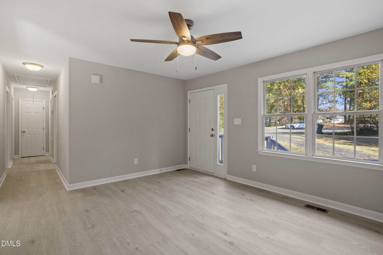 A bright, empty room with light wood flooring, beige walls, a ceiling fan with wooden blades, a white entrance door, and a large window showing a view of trees and a street outside.