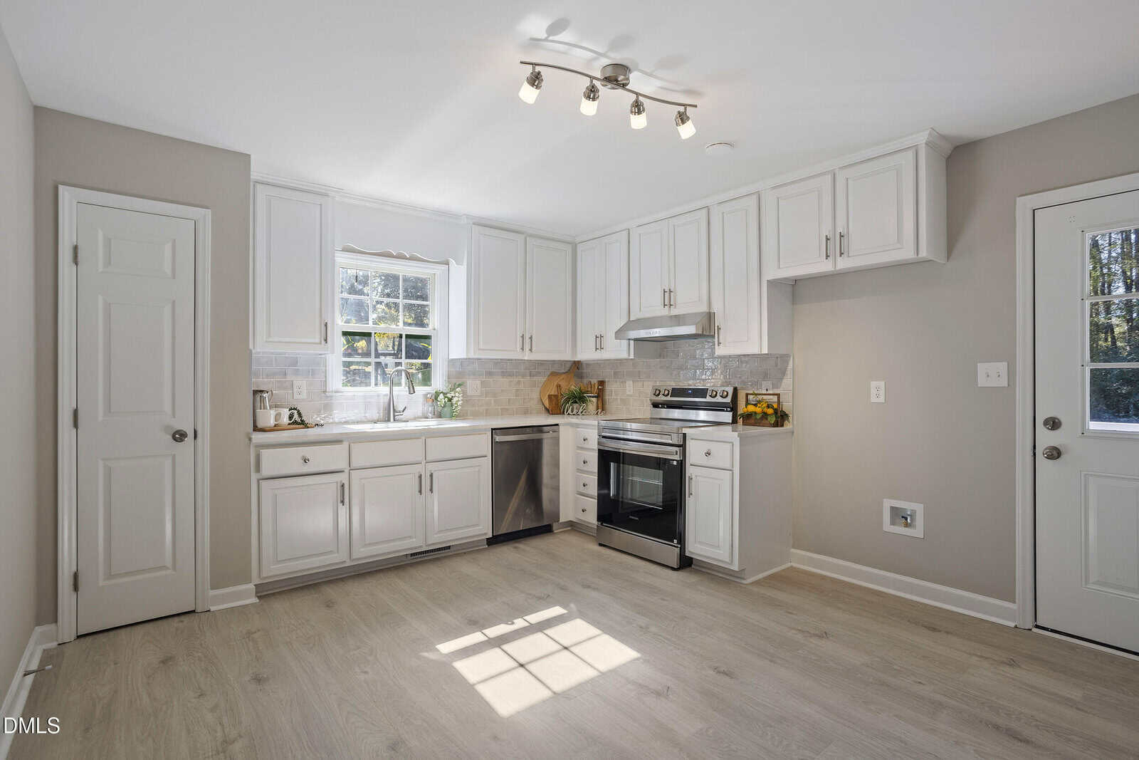 A modern kitchen with white cabinets, stainless steel appliances, light wood flooring, a window above the sink, and a door to the outside. The room is well-lit with ceiling spotlights and natural light.