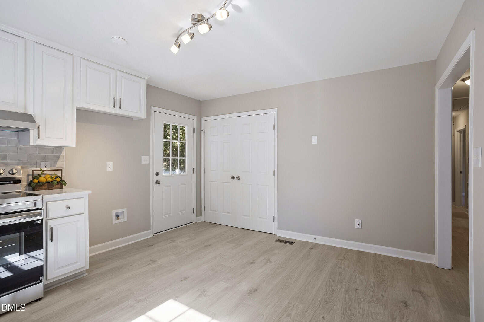 A modern kitchen with light wood flooring, white cabinets, stainless steel appliances, a door with a window, and a double-door closet. The walls are light beige, and the room is well-lit with ceiling lights.