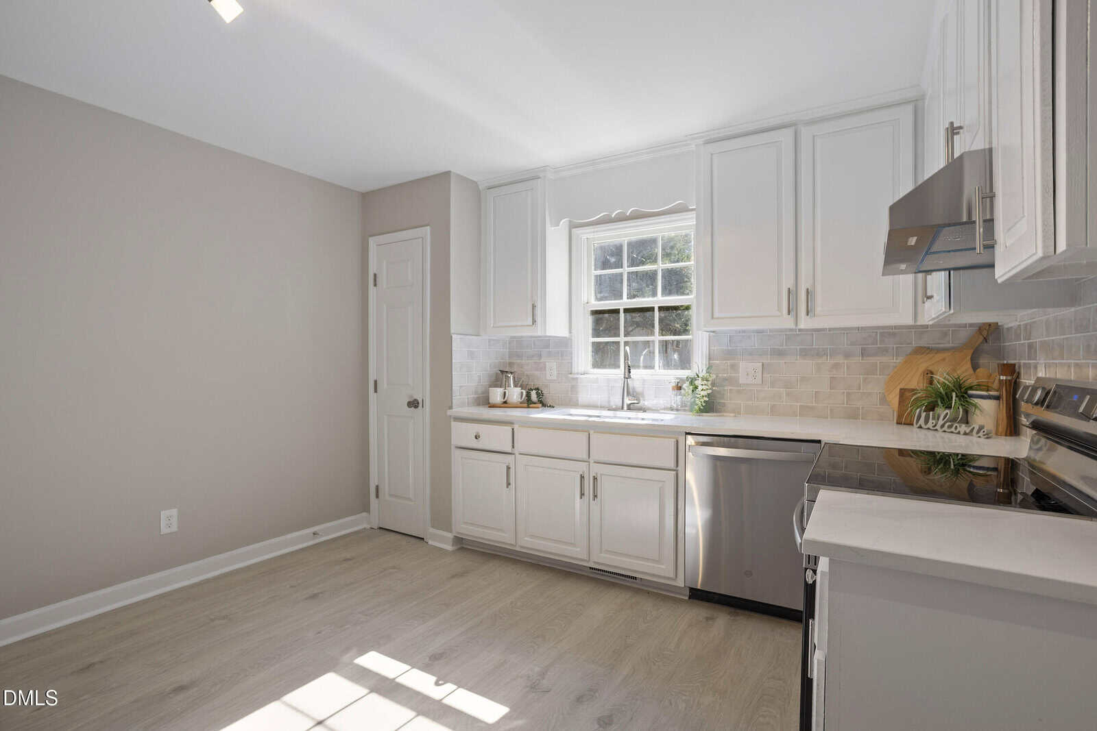 A modern kitchen with white cabinets, light wood flooring, stainless steel appliances, a window above the sink, and neutral-colored walls. A cutting board, utensils, and small plants are on the countertop.