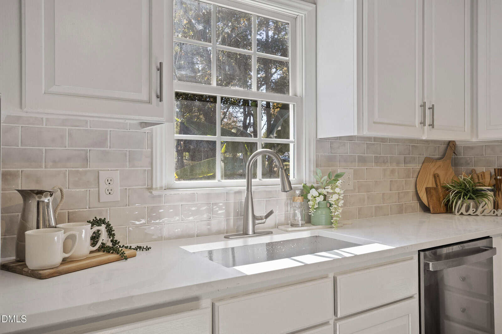 A bright kitchen with white cabinets and countertop, a stainless steel sink and faucet, light gray tile backsplash, and decorative items including cutting boards, a pitcher, greenery, and a "Welcome" sign. Sunlight comes through the window.