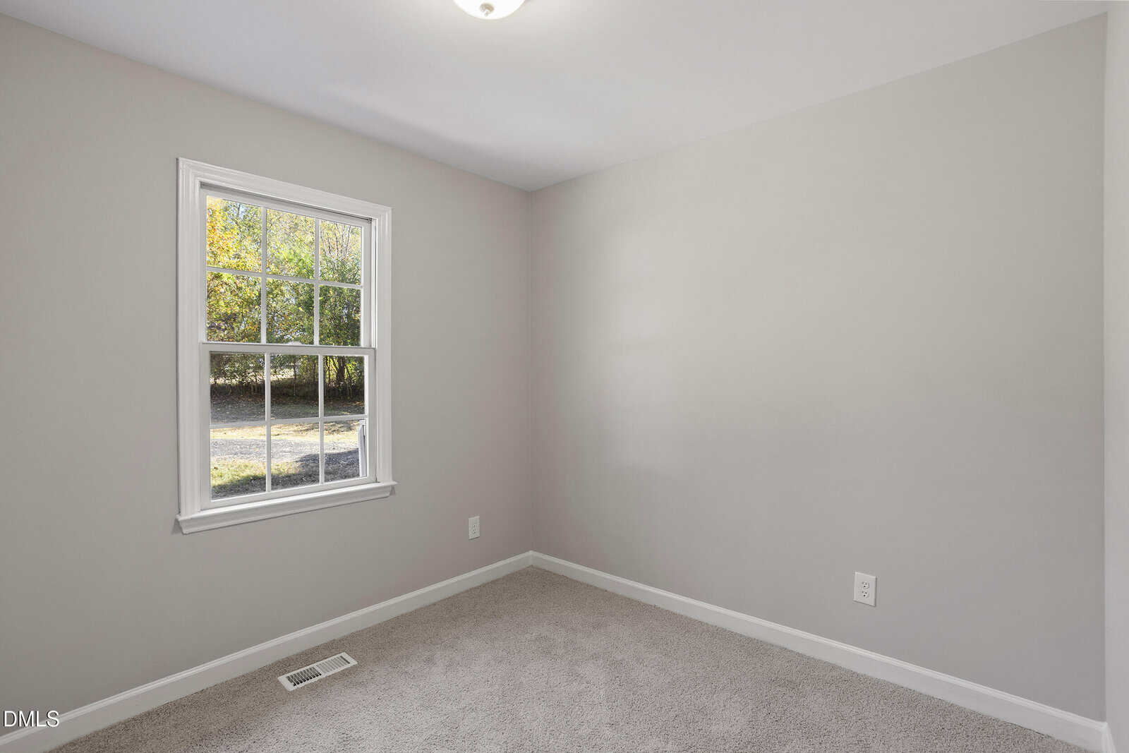An empty room with beige carpet, light gray walls, a white-framed window letting in natural light, and electrical outlets visible on the right wall. A vent is located on the floor beneath the window.