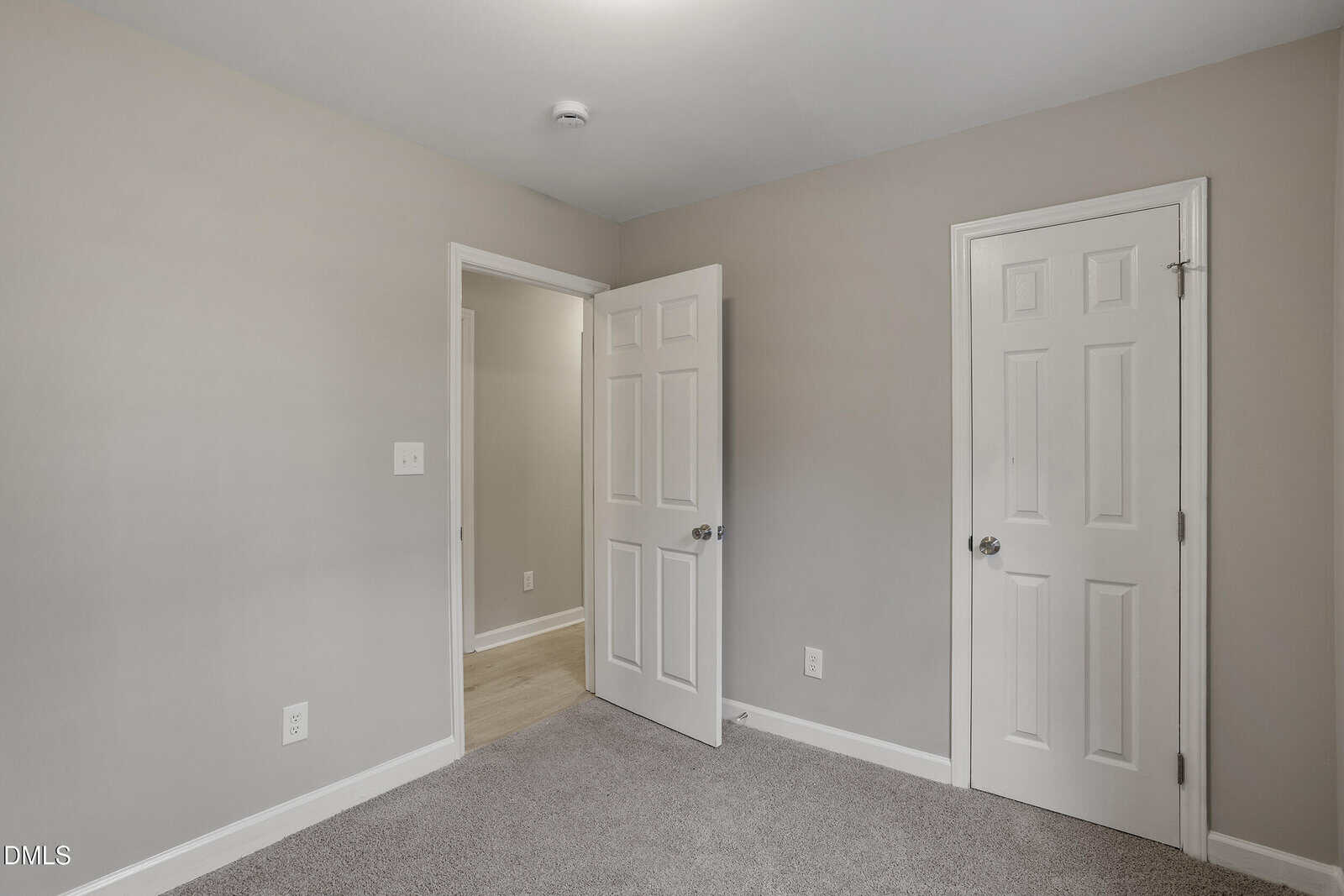 A small, empty room with light gray walls, beige carpet, white baseboards, two white doors, and a ceiling-mounted smoke detector. One door is slightly open, leading to a hallway with light wood flooring.