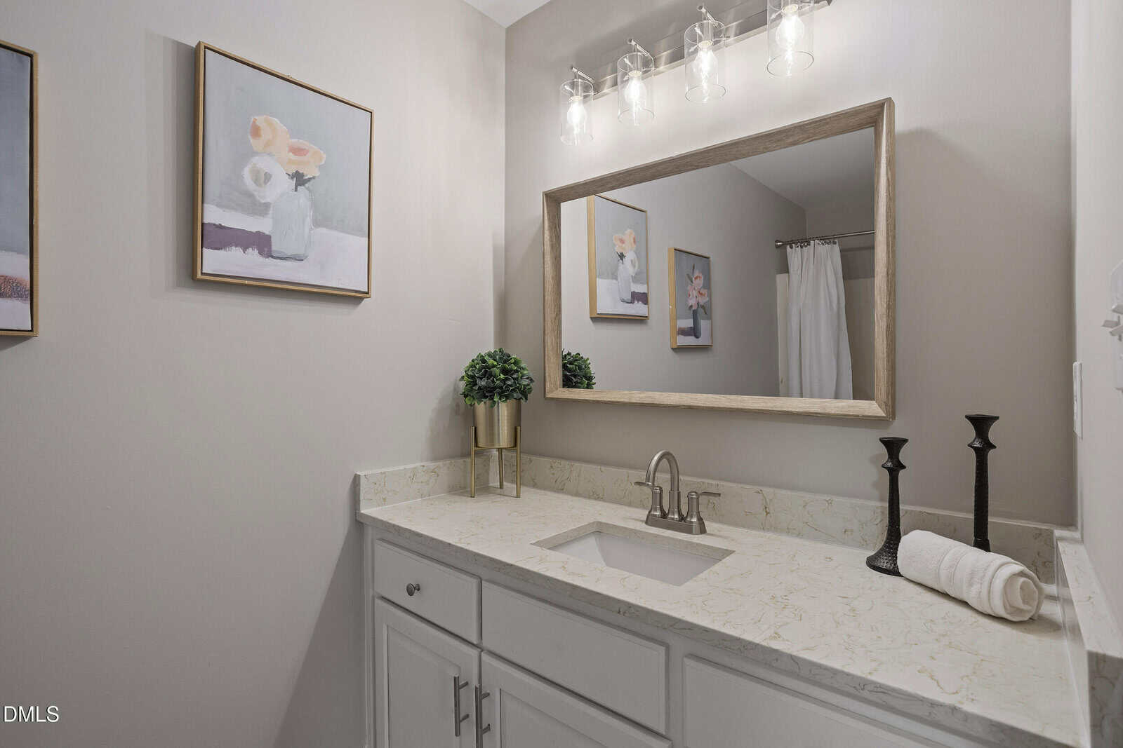 A bathroom with a marble countertop, rectangular mirror, sink, two black candle holders, a rolled towel, a small potted plant, and two floral paintings on light-colored walls.