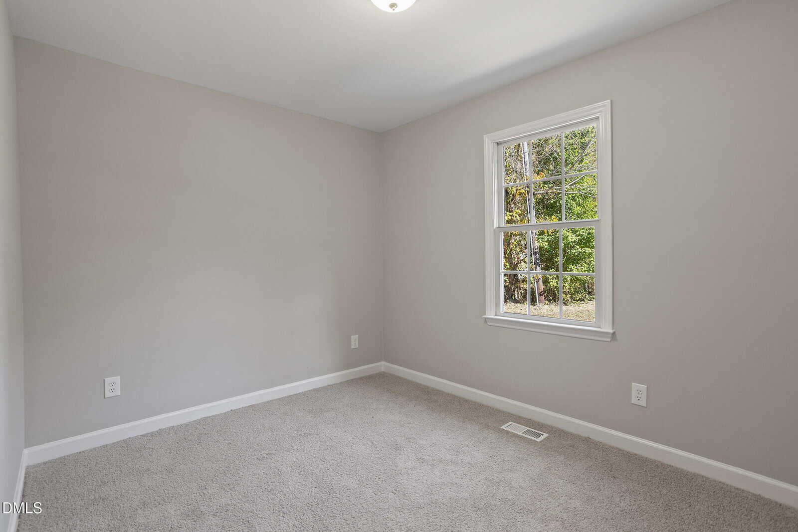 Empty room with beige carpet, light gray walls, white trim, a ceiling light, and a single window showing green trees outside. There are two electrical outlets and an air vent on the wall below the window.