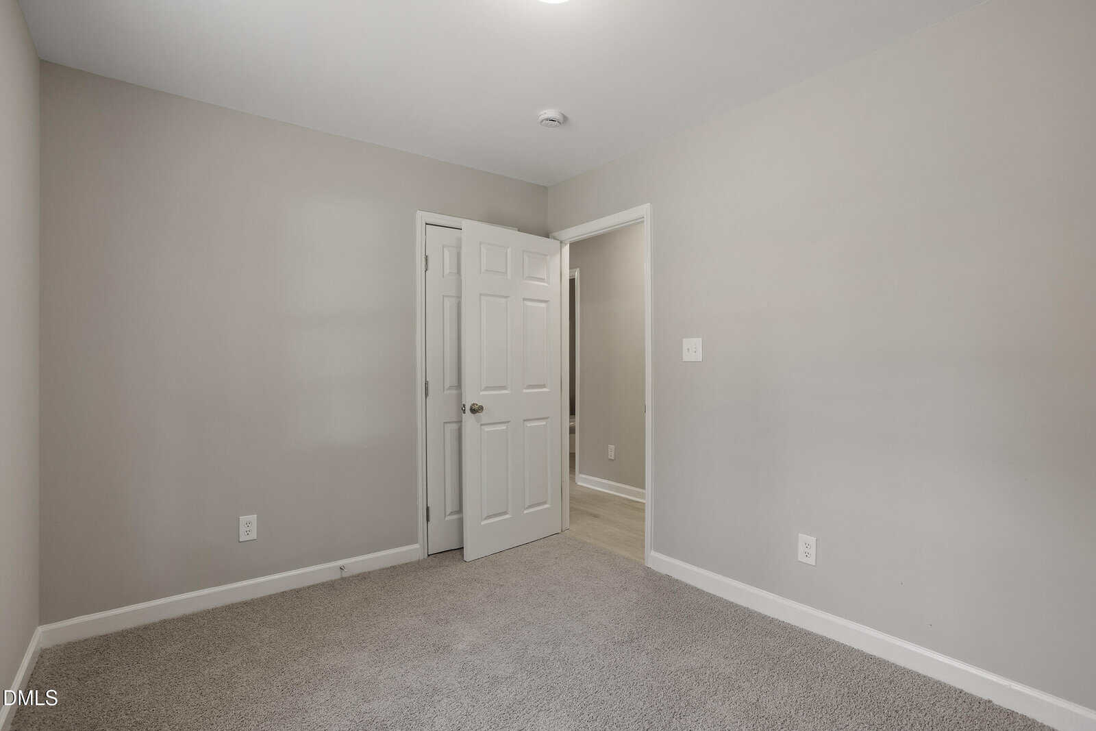 Empty room with beige walls, white trim, and carpeted floor. An open white door leads to a hallway with wood flooring. Ceiling light fixture is visible.
