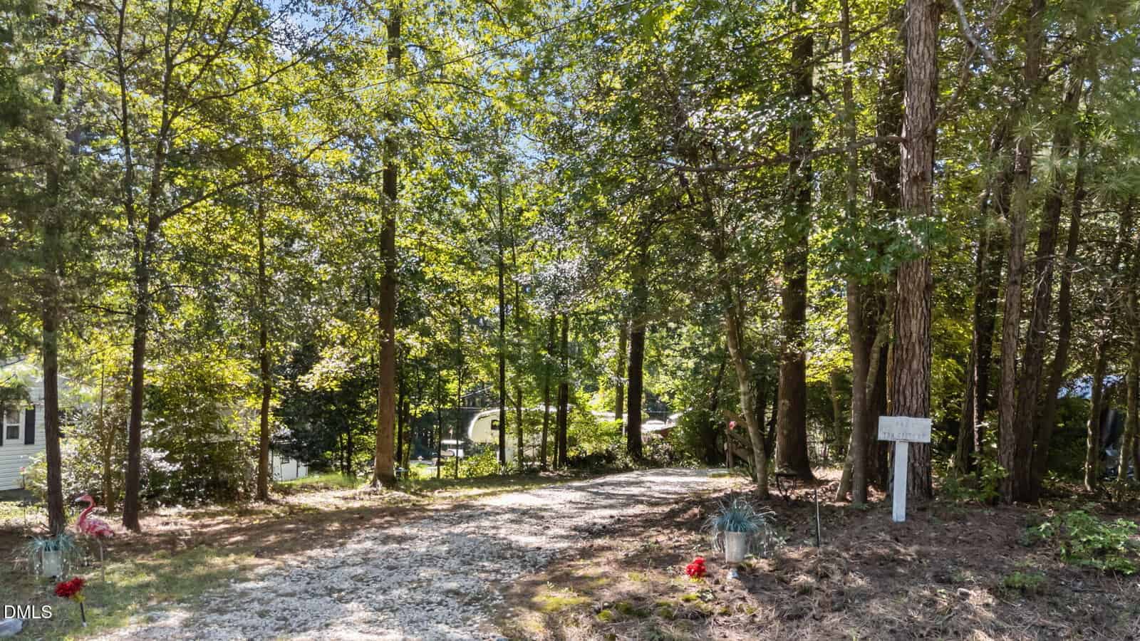 A gravel driveway at 136 Creek Drive, Louisburg, curves through a wooded area with tall trees and green foliage. Red flowers and a flamingo lawn ornament accent the left side, while a small white sign stands on the right by the trees.