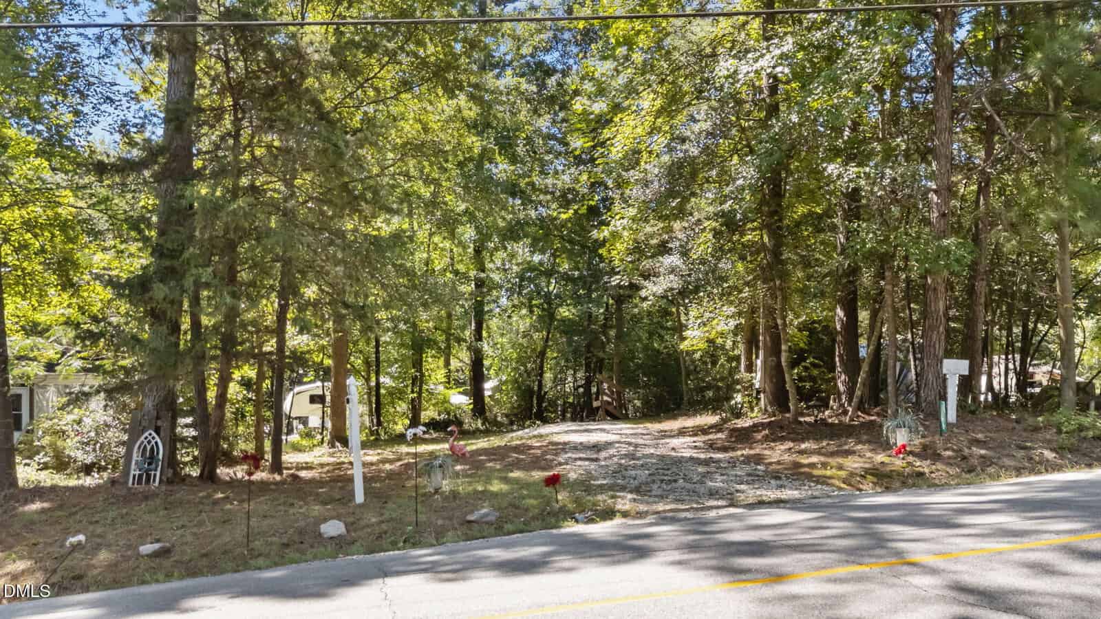 A gravel driveway leads from a paved road into a wooded area with tall green trees. There are mailboxes and some small decorations near the entrance, and sunlight filters through the foliage.