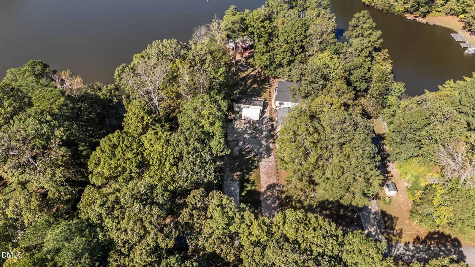Aerial view of a wooded lakeside property with a few buildings, a dirt driveway, and surrounding trees, bordered by water on two sides, under a clear sky.
