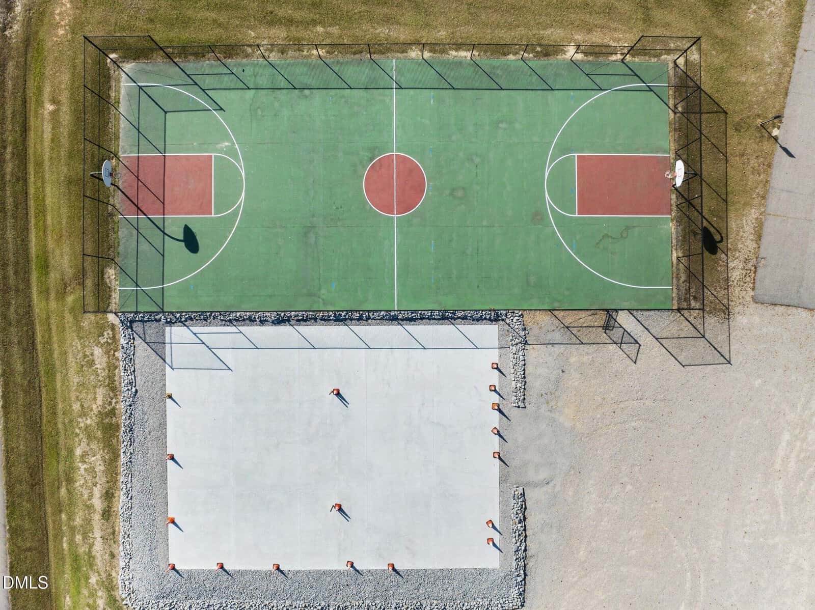 Aerial view of an outdoor basketball court with green and red markings, next to a fenced, rectangular concrete area with poles casting shadows. Grass and gravel surround both surfaces.