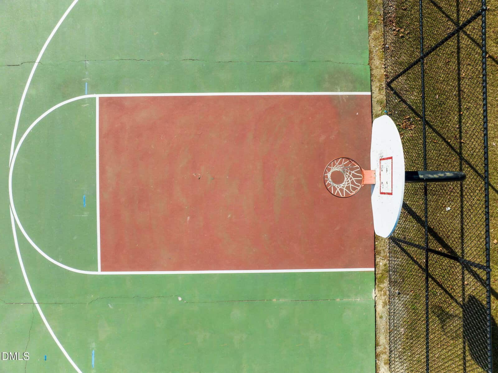 Aerial view of an outdoor basketball court showing the hoop, backboard, and painted key area with green and red surface. A black fence runs along the lower right edge of the court.
