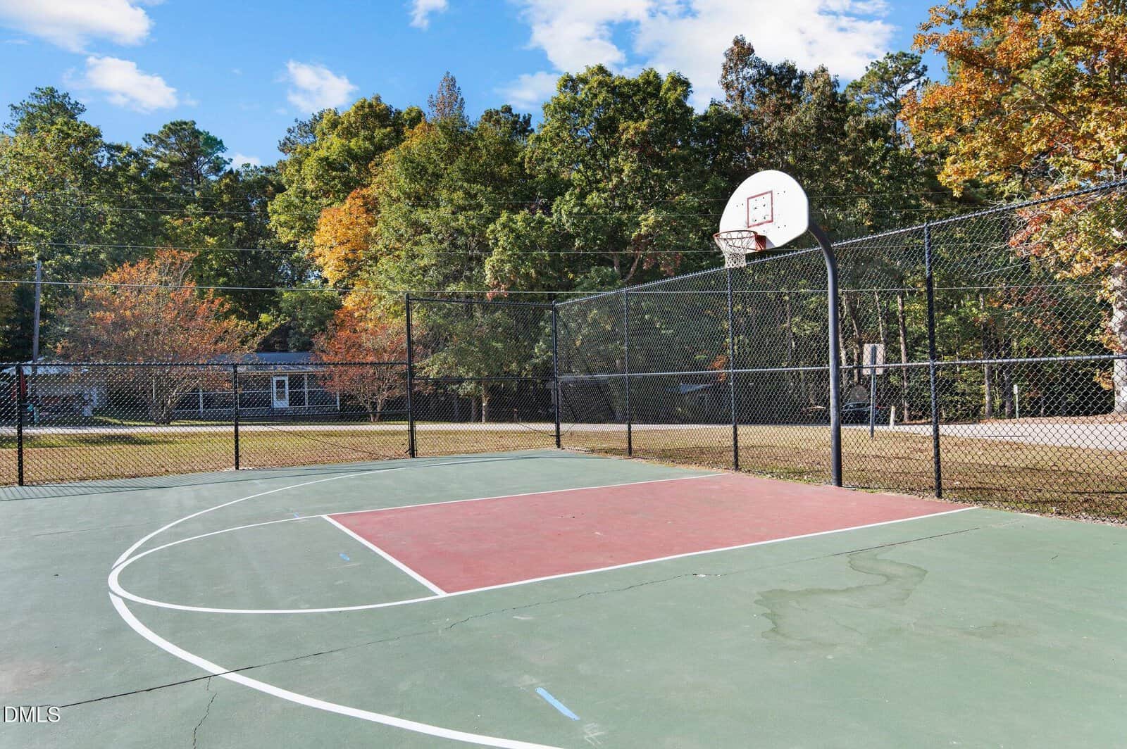 An outdoor basketball court with a red-painted key, surrounded by a black chain-link fence, trees with autumn foliage, and a house in the background under a partly cloudy sky.
