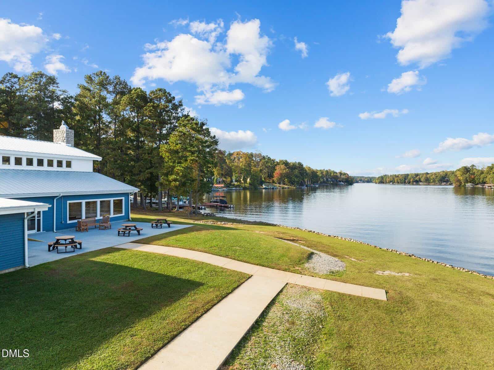 A lakeside scene with a blue building, picnic tables on grassy areas, trees along the shore, and a calm lake under a blue sky with scattered clouds. A paved walkway leads toward the water.