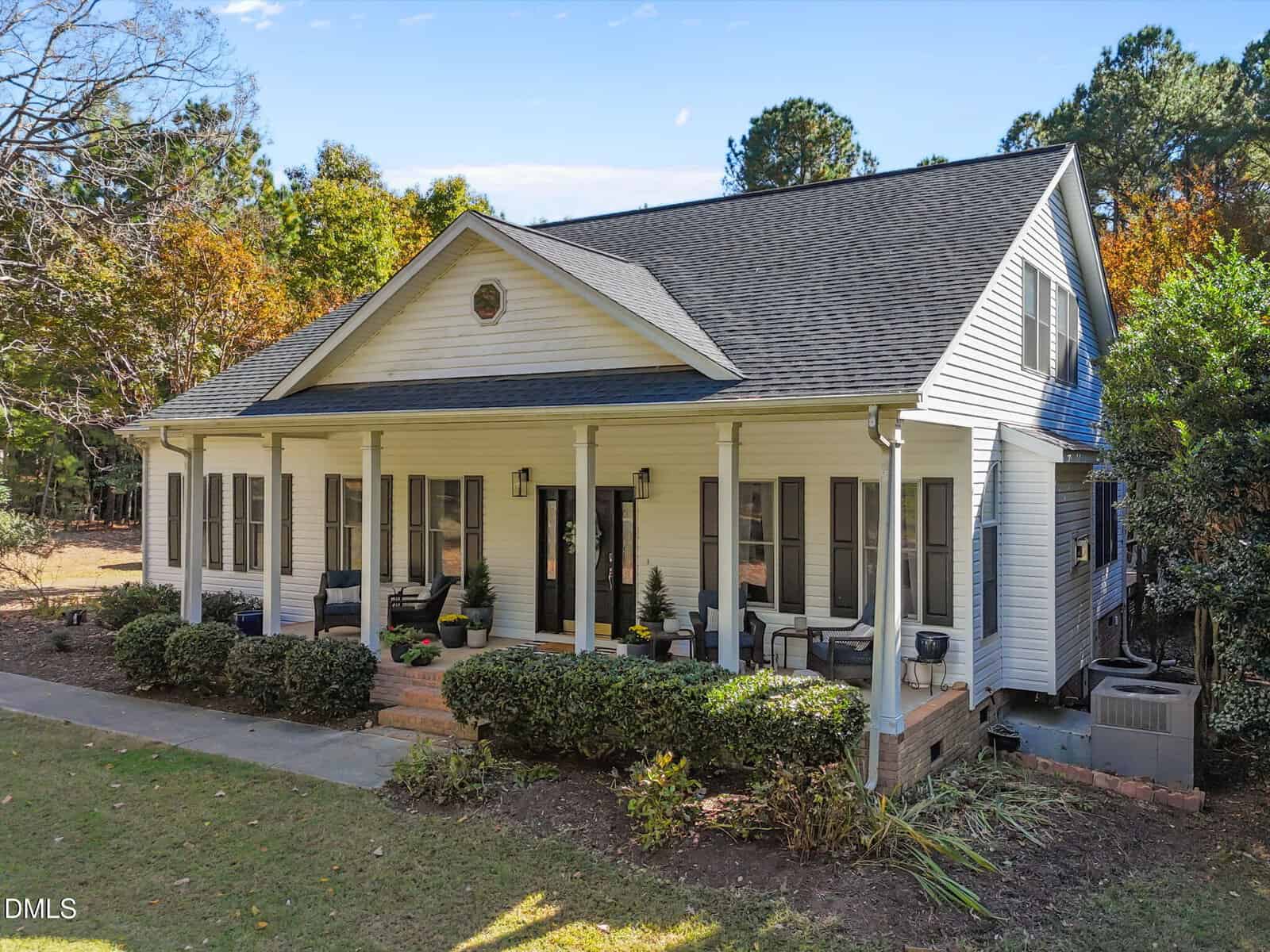 A white house with a large covered porch supported by columns, black shutters, and a dark shingle roof. The porch has several chairs and potted plants. Shrubs and grass line the front yard. Trees are visible in the background.
