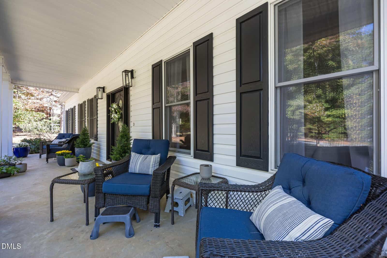 A covered porch with white siding, black shutters, blue cushioned chairs, potted plants, and a small side table. Trees and greenery are visible in the background. The setting is clean and tidy.
