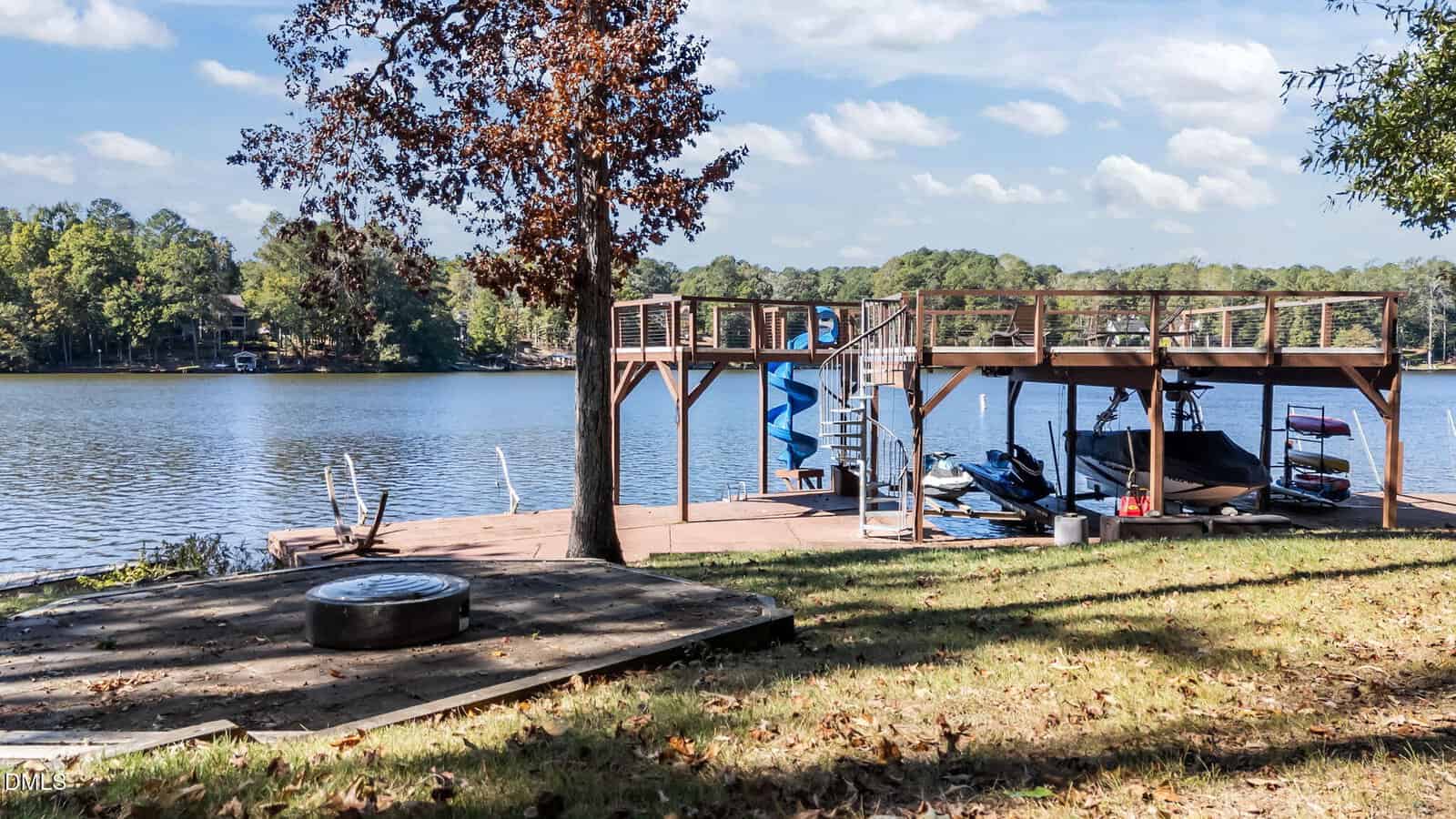 A wooden dock with a covered boat slip, a jet ski, and a blue spiral slide sits on a peaceful lake, surrounded by trees. A grassy yard and a circular fire pit are in the foreground.