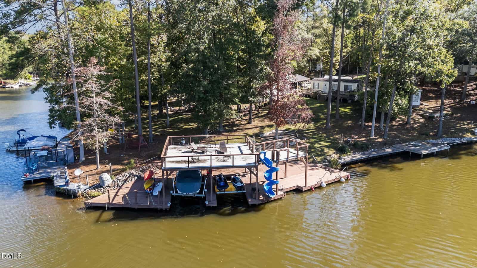 A wooden dock with a covered seating area, boats, kayaks, and a blue spiral slide sits on a tree-lined lakeshore, with houses and forest visible in the background.