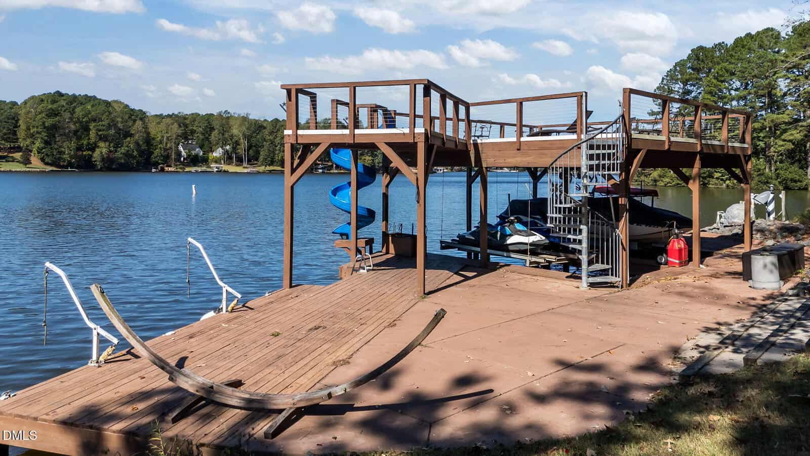 A wooden dock with a spiral staircase, blue slide, and covered boat area extends over a calm lake. An empty hammock stand sits in the foreground, surrounded by trees and grassy shoreline under a sunny sky.