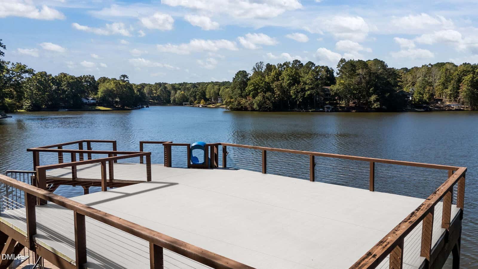 A wooden dock with railings extends over a calm lake, surrounded by trees under a partly cloudy sky. A blue slide is attached to the dock, and the water reflects the greenery and sky.