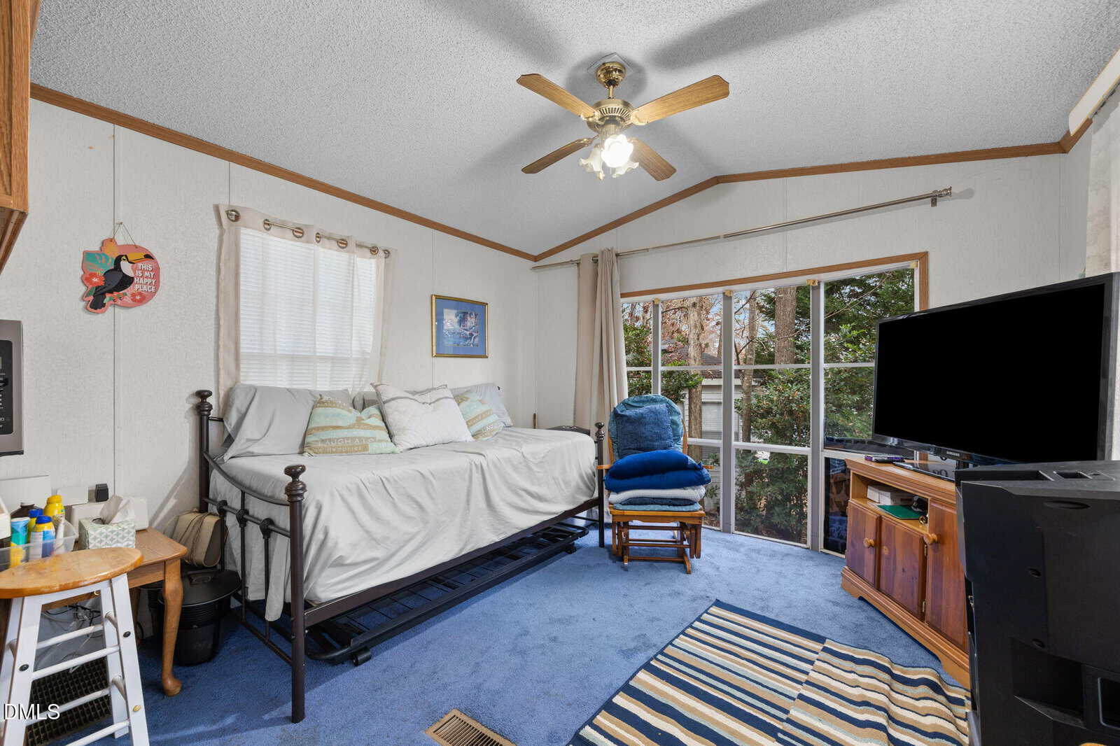A cozy bedroom with blue carpet, a black metal daybed, a blue armchair, and a wooden TV stand in front of large windows that let in natural light. A ceiling fan is above, and various decor items are on the walls and shelves.