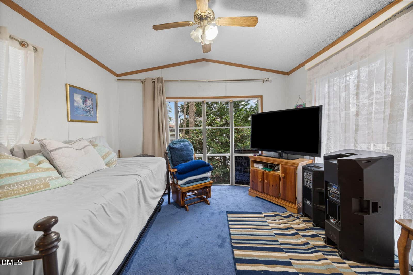Bright cozy room with blue carpet, daybed with pillows, armchair, TV on wooden stand, speakers, striped rug, large windows with sheer curtains, ceiling fan, and framed wall art.