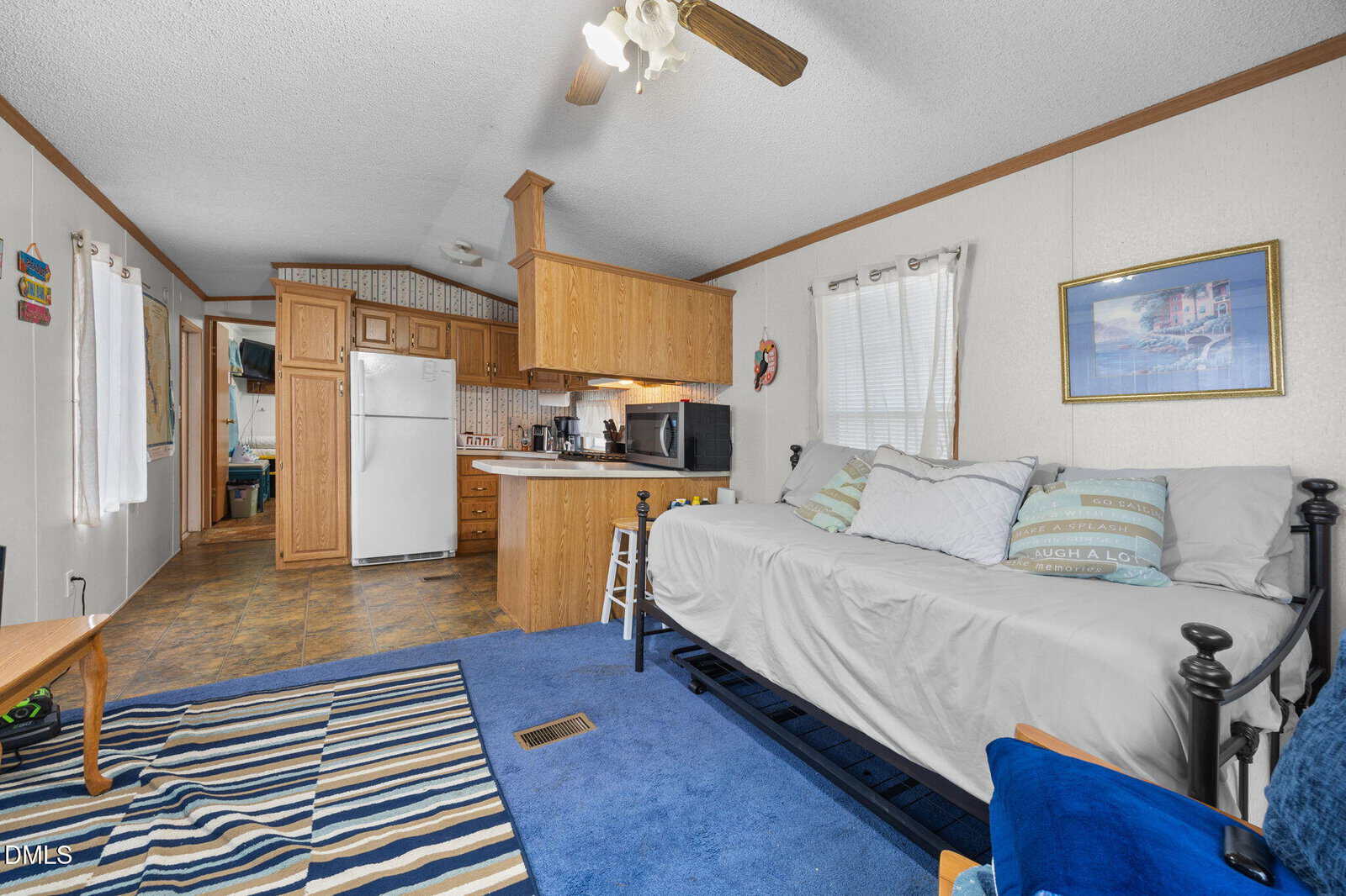 A cozy living area with a daybed, blue-striped rug, small kitchen with wooden cabinets, and white appliances. Light streams in from a window with sheer curtains. Coastal-themed decor accents the space.