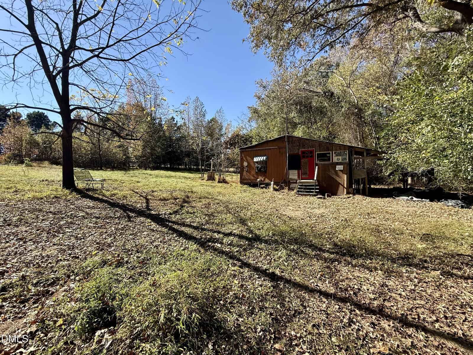 A small wooden cabin with a porch and signs on the front sits in a grassy, partially shaded clearing surrounded by trees under a clear blue sky. Fallen leaves cover the ground in the foreground.