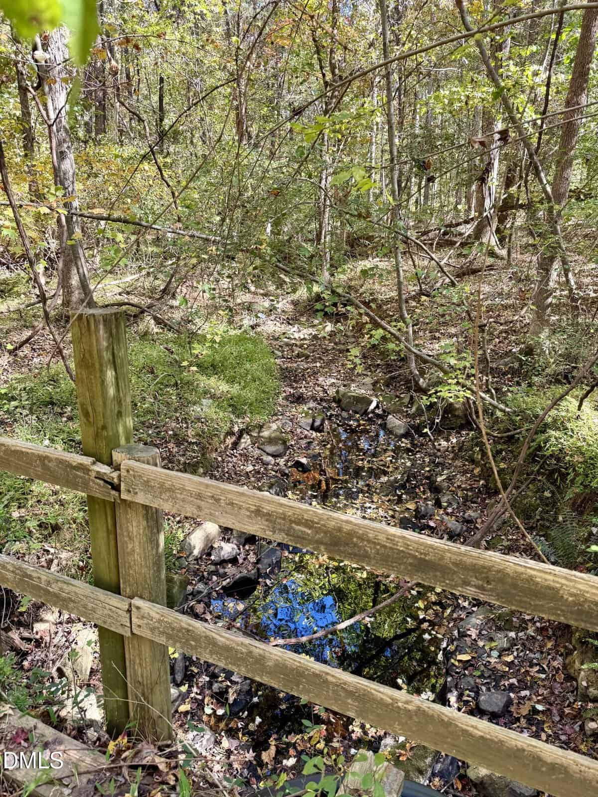 A wooden fence in the foreground runs alongside a small creek with rocks and water, surrounded by green foliage and trees in a wooded area.