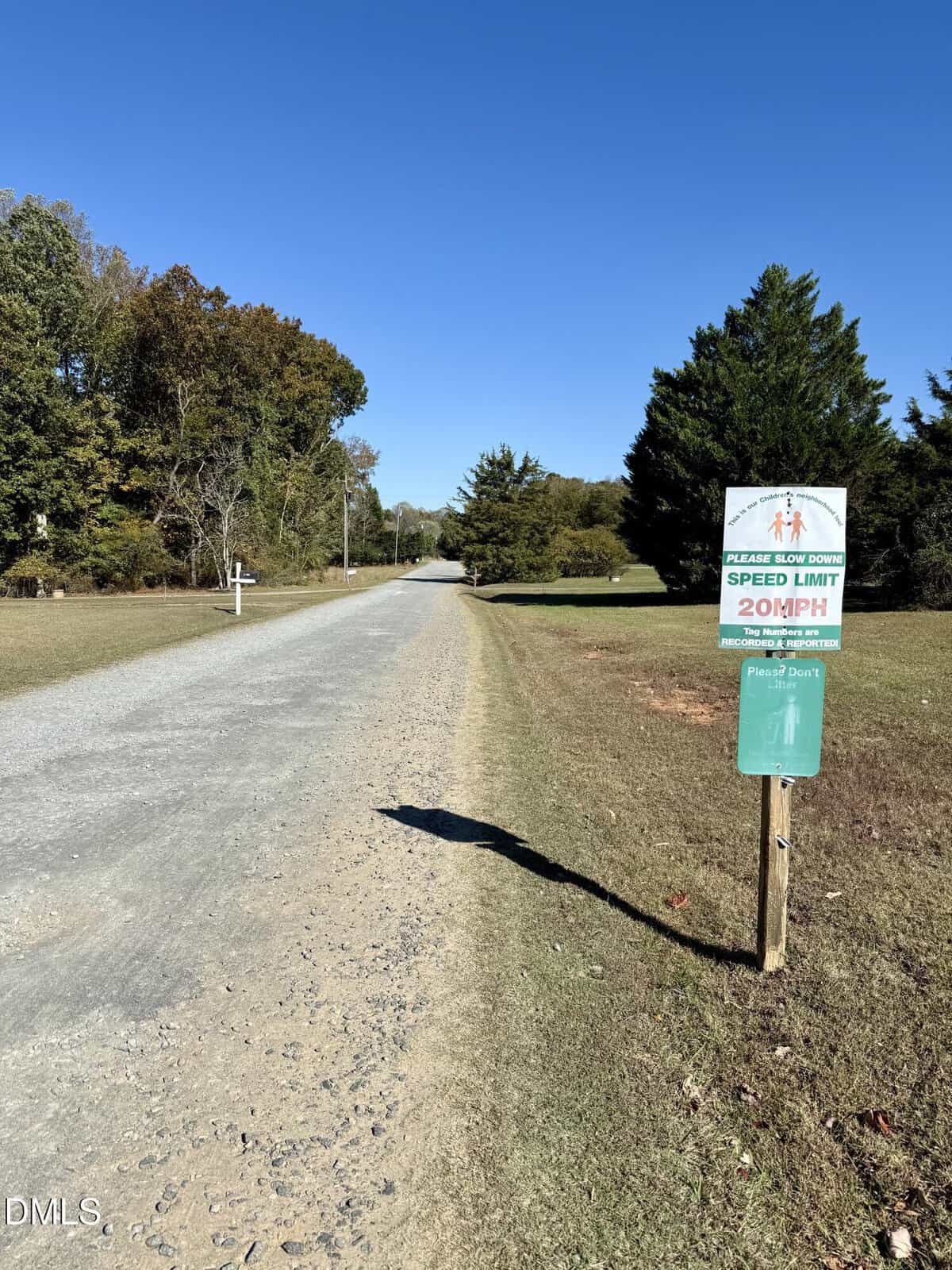 A gravel road runs through a rural area bordered by grass and trees. A sign on the right side of the road indicates a speed limit of 20 MPH. The sky is clear and blue.