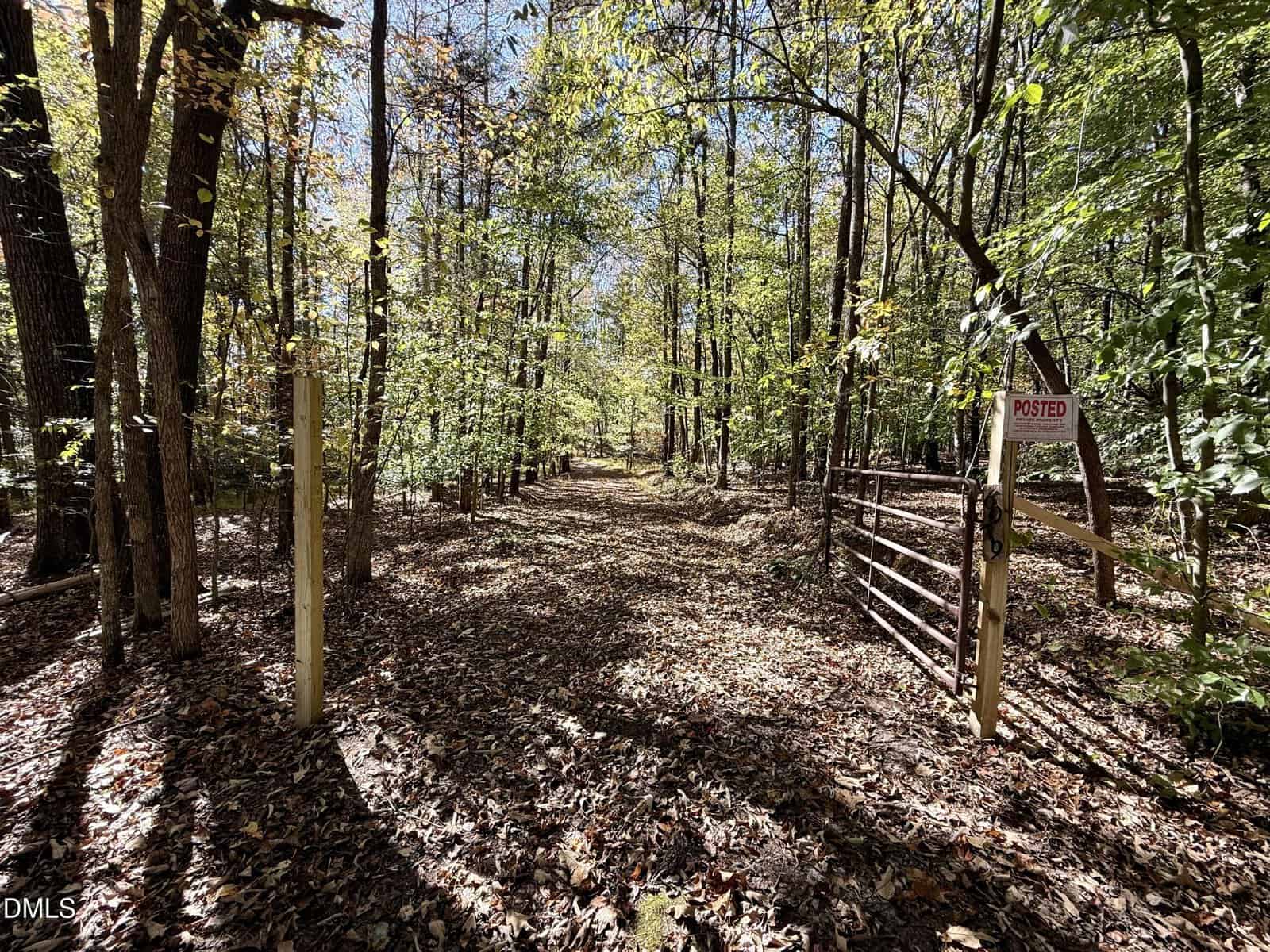 A dirt path covered with fallen leaves runs through a wooded area with tall trees. A metal gate and "Posted" sign are on the right side of the entrance, with sunlight filtering through the foliage.