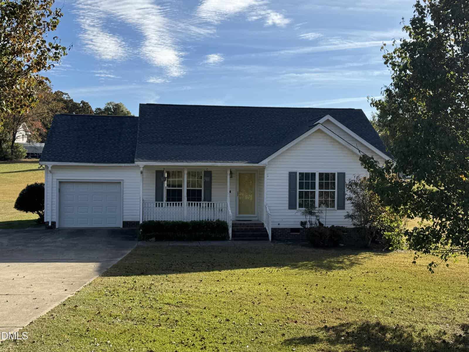 1318 Bobbitt Road in Kittrell is a single-story white house with a covered front porch, attached one-car garage, and dark roof. It sits on a grassy yard with trees and a driveway, beneath a partly cloudy sky.