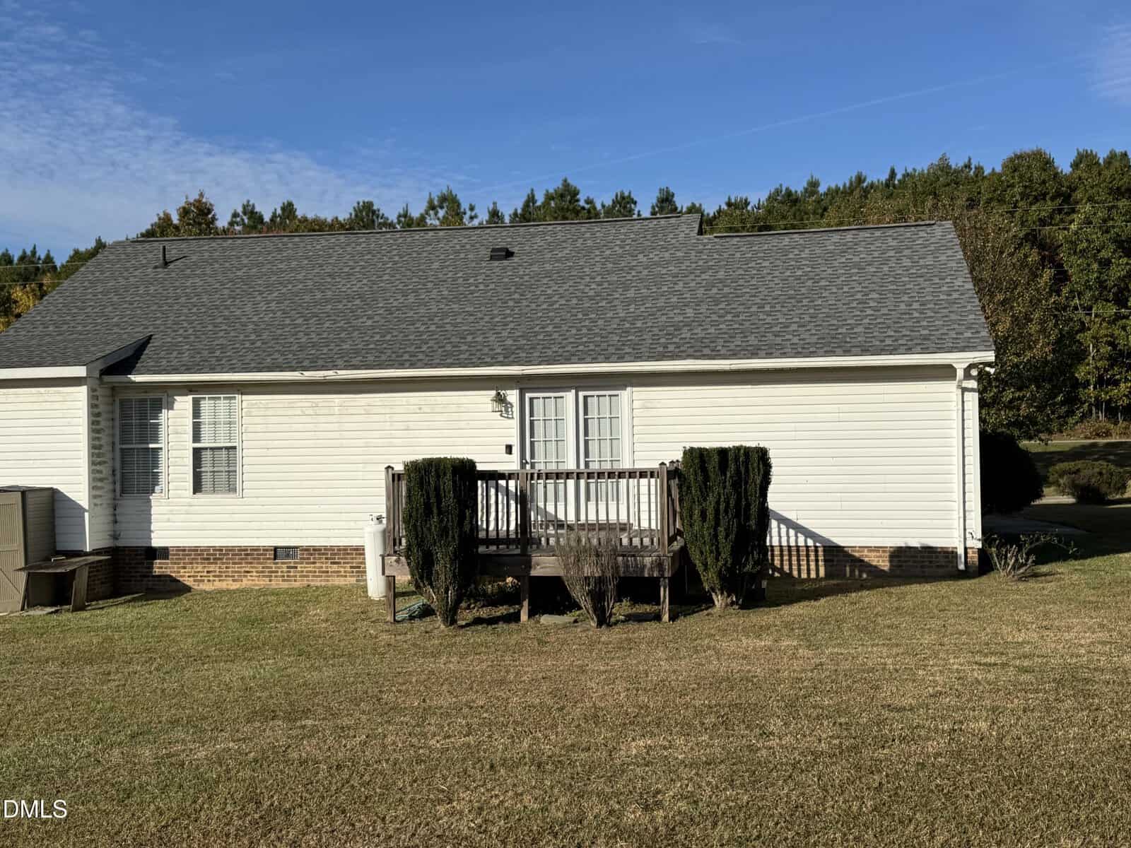 A small wooden porch with steps and trimmed shrubs in front of a one-story white house with gray shingles, brick foundation, and a grassy yard. Trees are visible in the background under a blue sky.