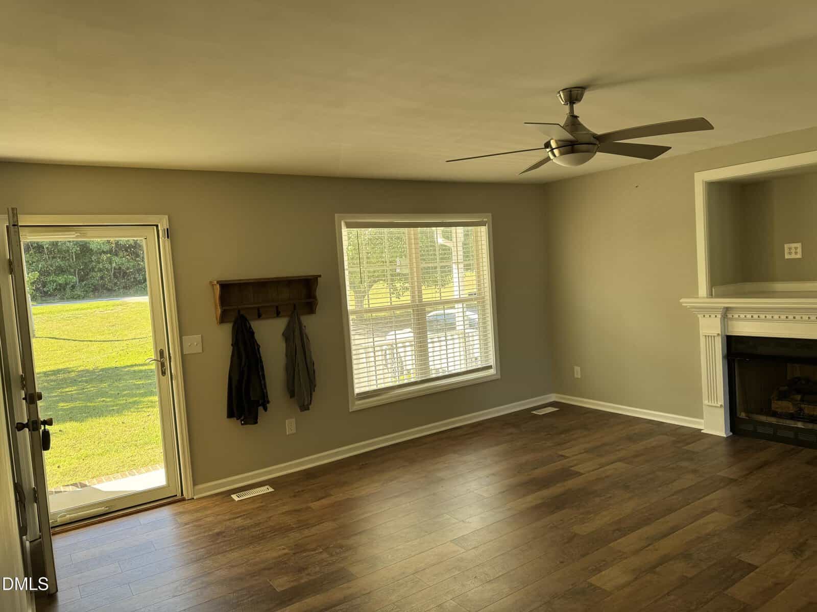 A living room with hardwood floors, a ceiling fan, a large window with blinds, an open front door, a wall-mounted coat rack with jackets, and a fireplace with a white mantel.