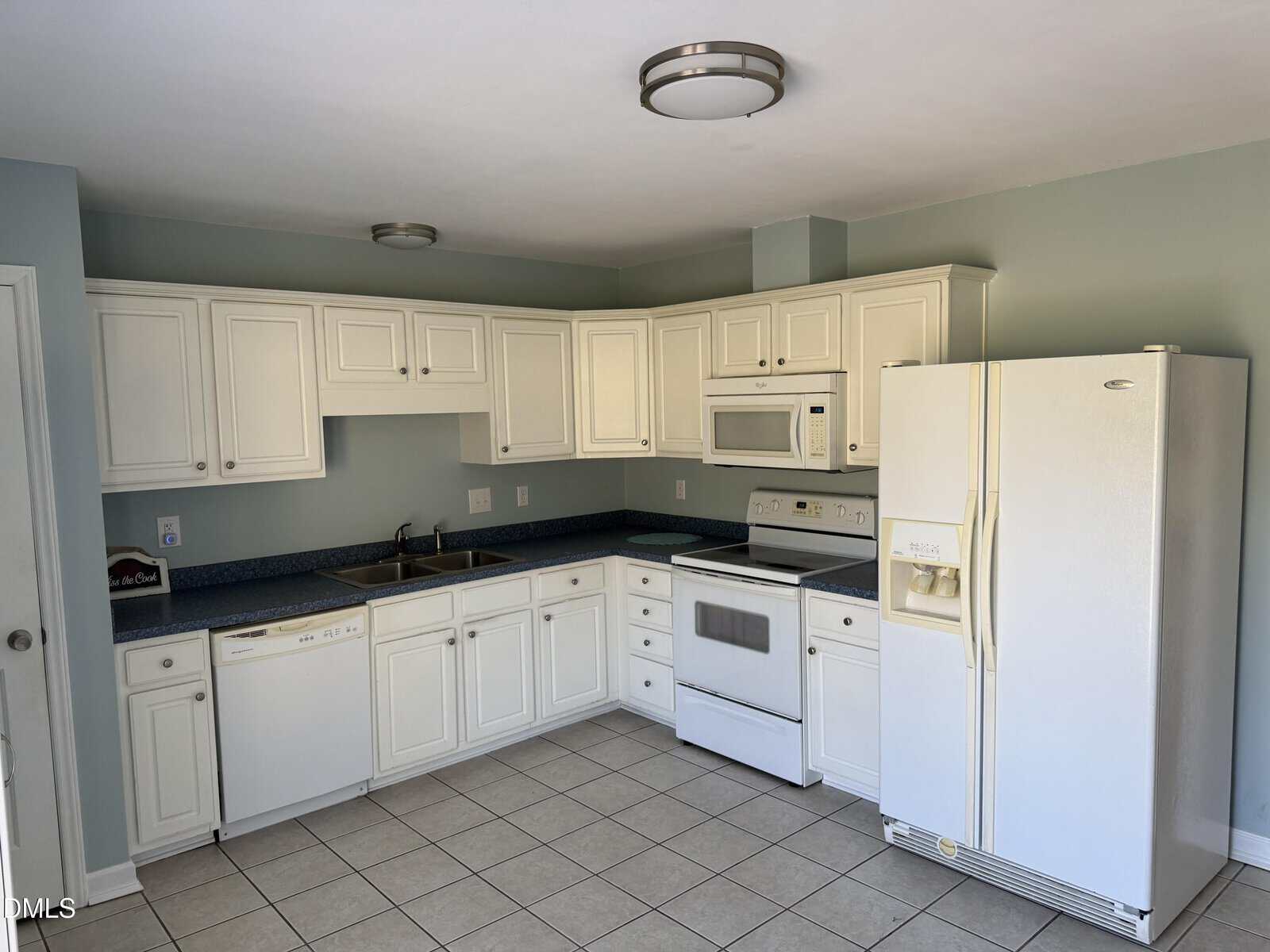 A kitchen with white cabinets, white appliances including a refrigerator, stove, dishwasher, and microwave, blue countertops, a double sink, tile flooring, and pale blue walls.
