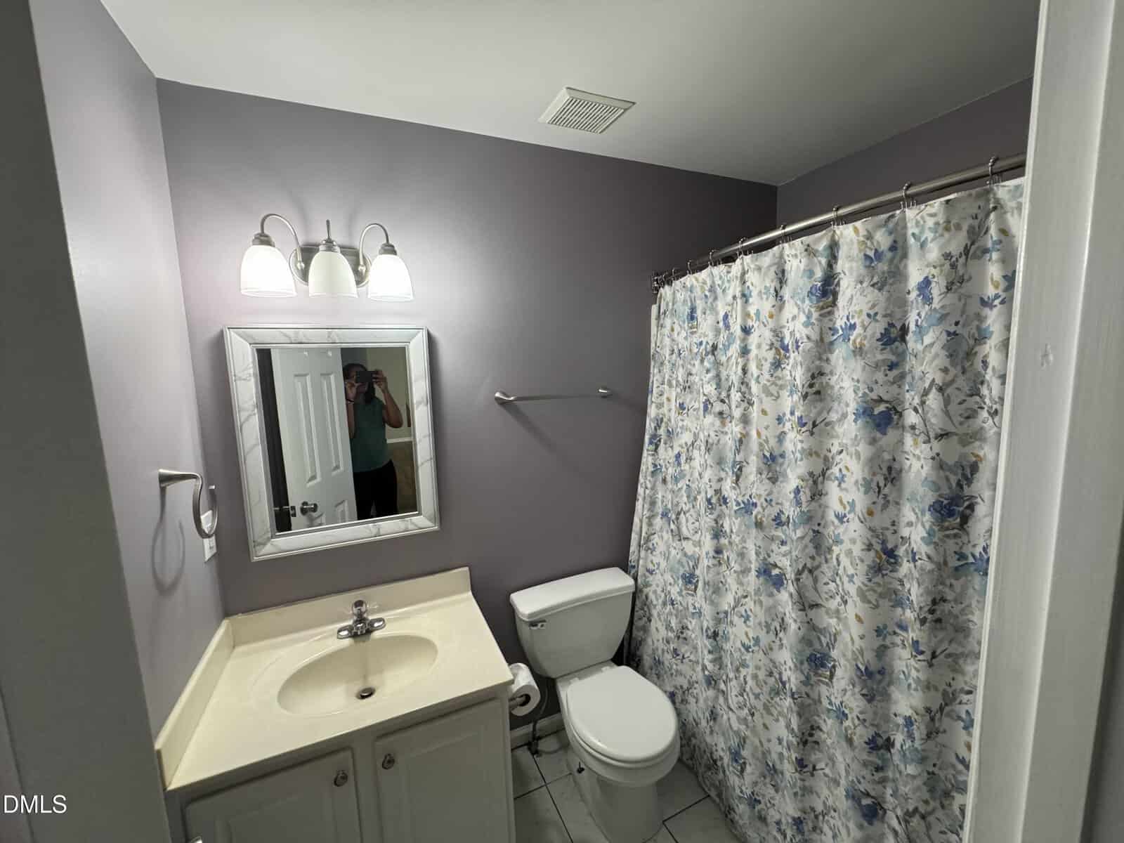 Small bathroom with light purple walls, a white sink and toilet, a mirror, and a shower with a blue floral curtain. Three lights are above the mirror, and a person is visible in the mirror taking the photo.
