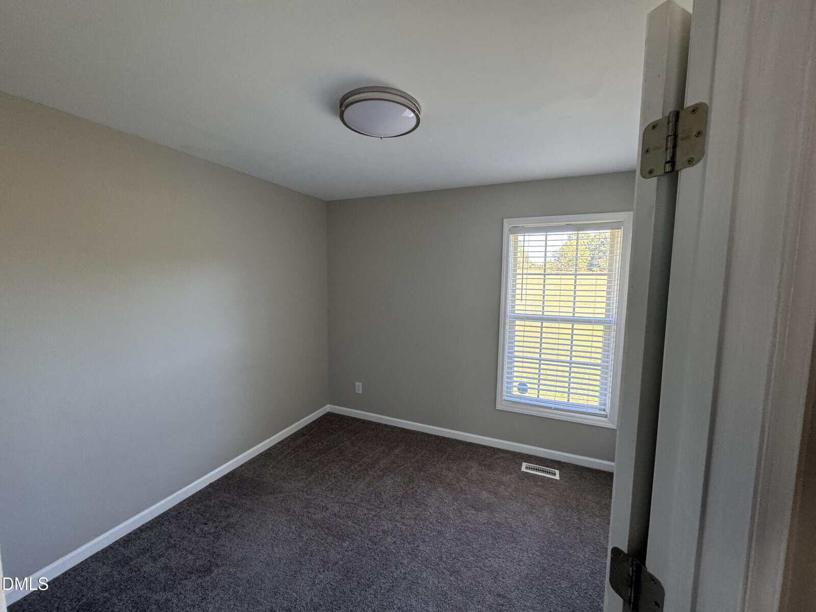 Small empty room with gray carpeting, light gray walls, a ceiling light, and a single window with blinds letting in natural light. Door is partially open, showing the simple and clean interior.