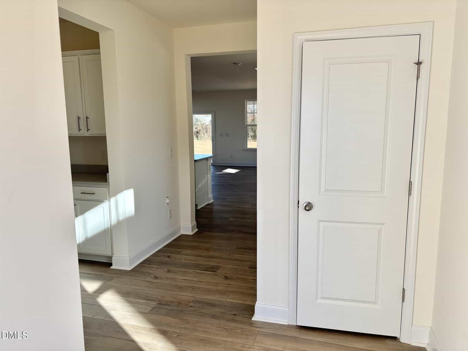 A bright, empty hallway with light wood floors, white walls, a closed white door, and openings leading to a kitchen area and a sunlit room in the background.