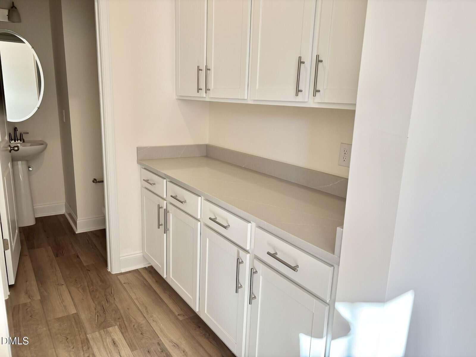 A bright hallway features white cabinets and drawers with silver handles, a gray countertop, and light wood flooring. An adjacent room with a round mirror and pedestal sink is partially visible.