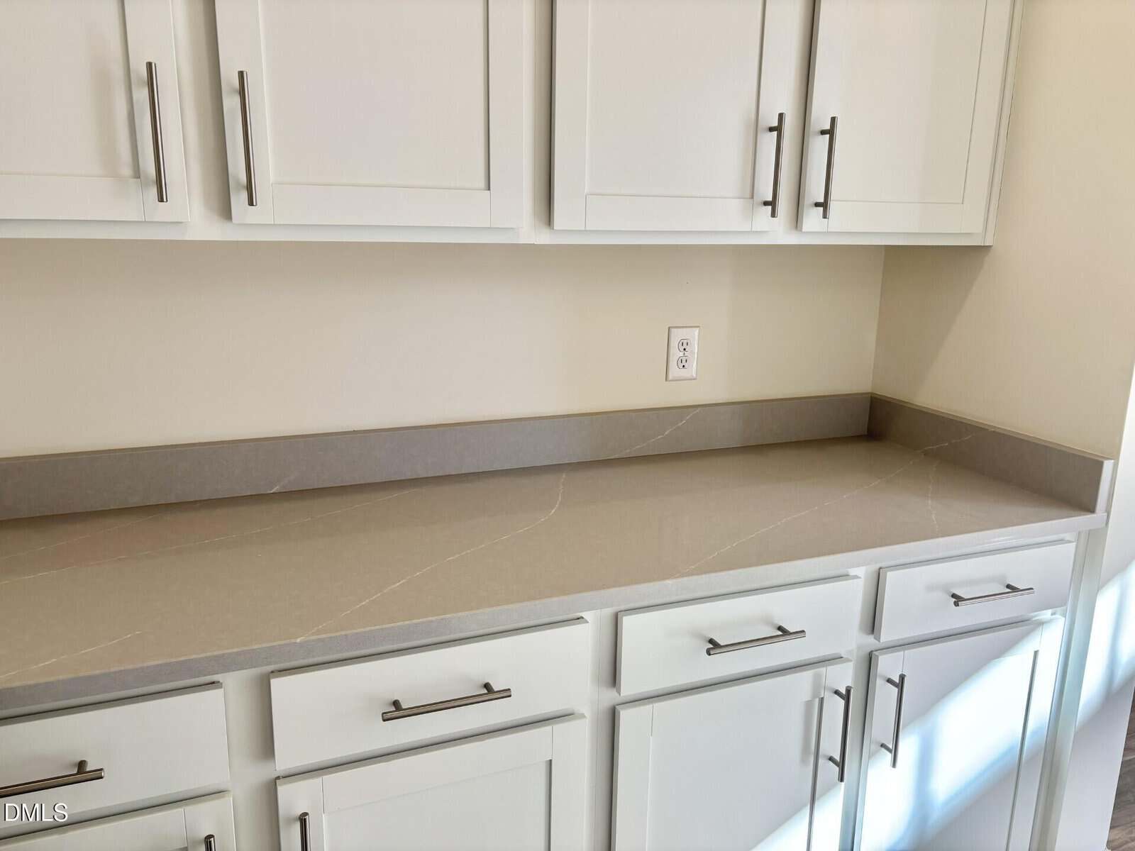 A kitchen countertop with light gray stone surface, white cabinets and drawers beneath, and matching upper cabinets. All cabinets have modern silver handles; there is an electrical outlet on the wall above the counter.