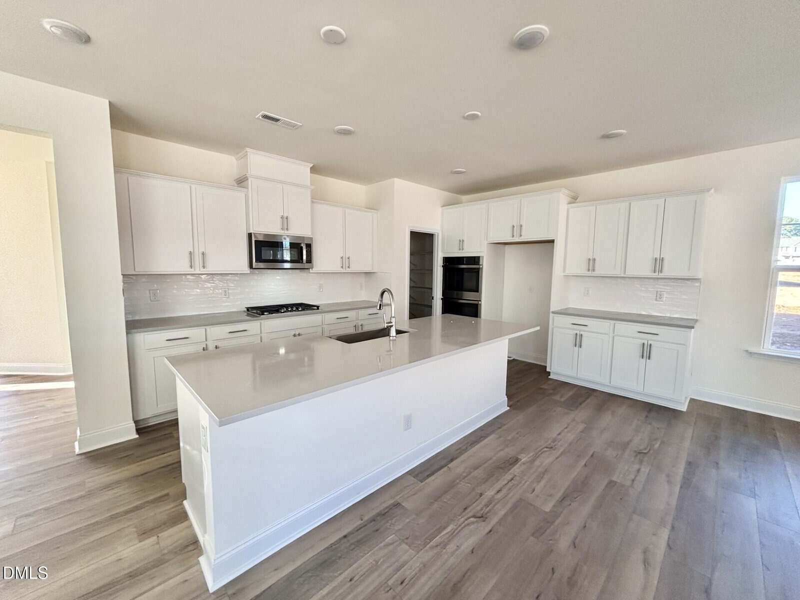 Modern kitchen with white cabinets, stainless steel appliances, a large island with a sink, light gray countertops, wood-style flooring, and bright natural light from windows.