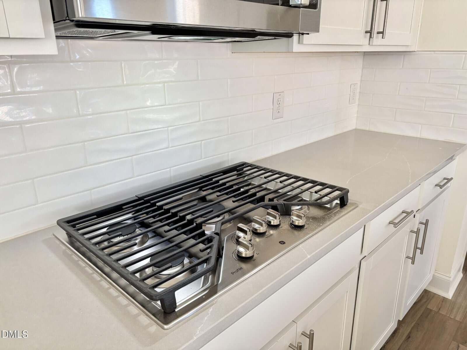 Modern kitchen countertop with a stainless steel gas cooktop, four control knobs, white cabinets, subway tile backsplash, and wood flooring.